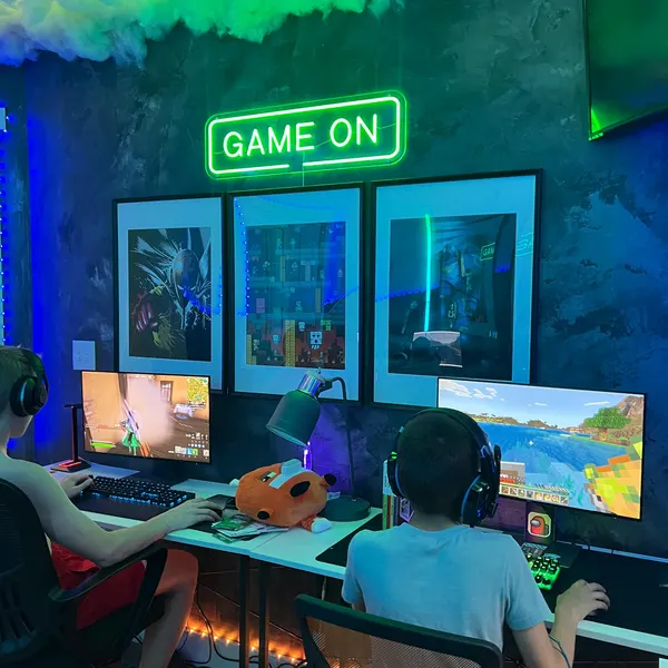 Green 'Game On' LED neon sign above two kids playing on dual monitors in a framed-art gaming den