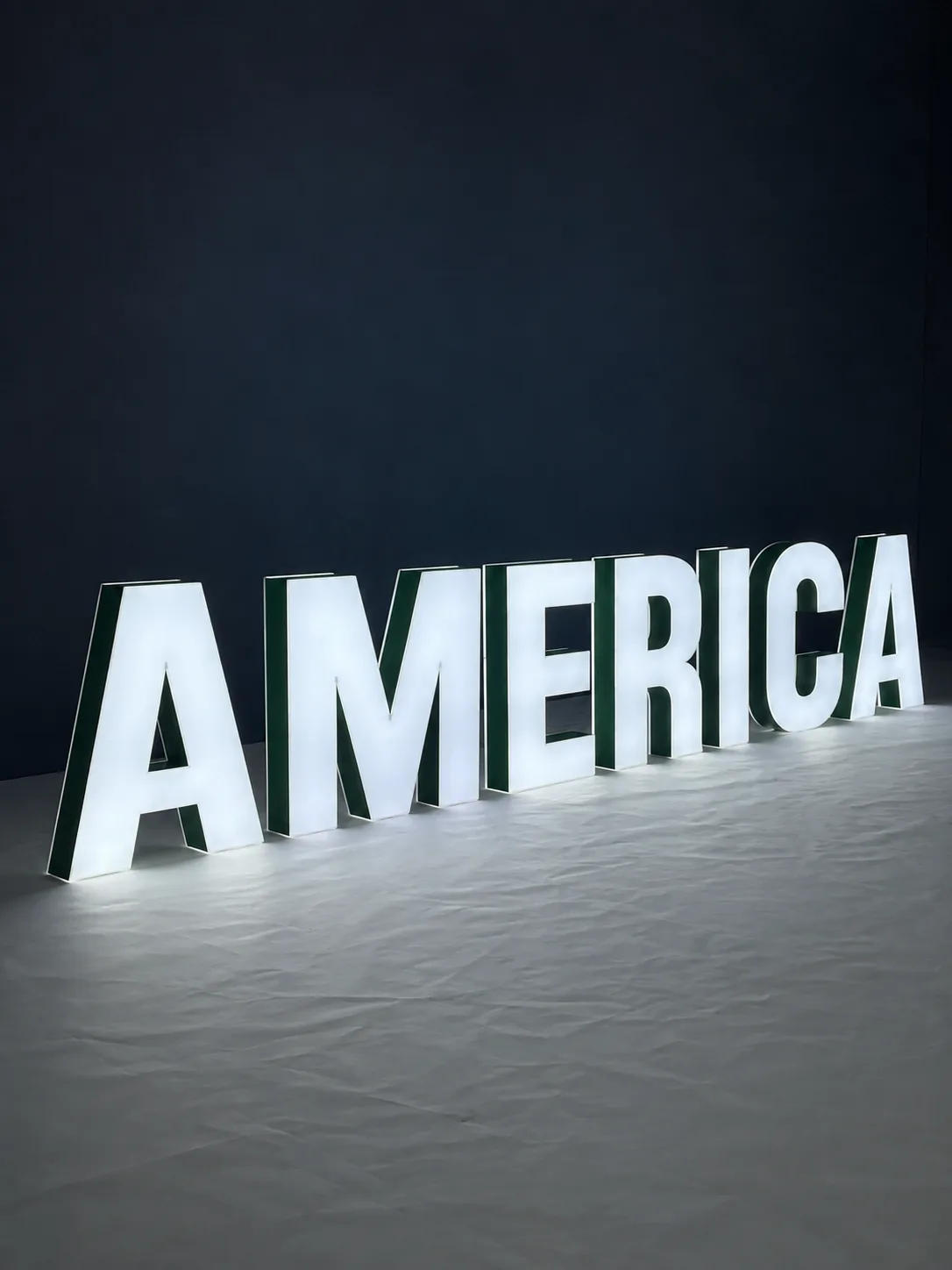 Dual-lit 3D metal letters spelling AMERICA, with glowing white faces and a halo of light spilling onto the floor behind