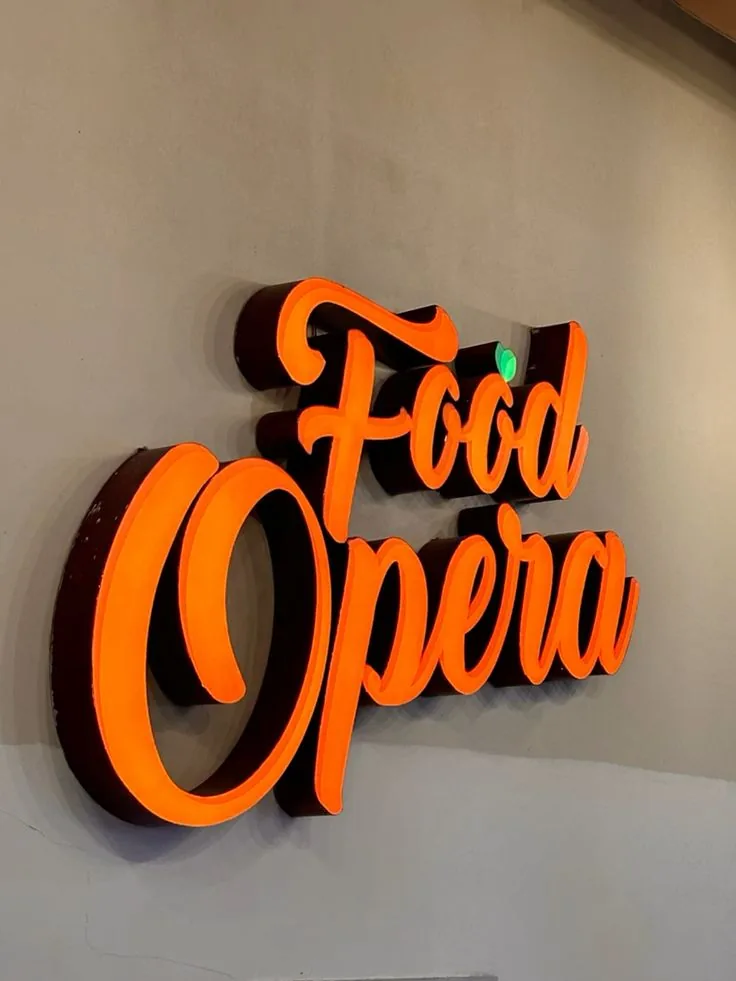 Frontlit 3D metal channel letters spelling Food Opera, the script faces glowing bright orange against a plain wall